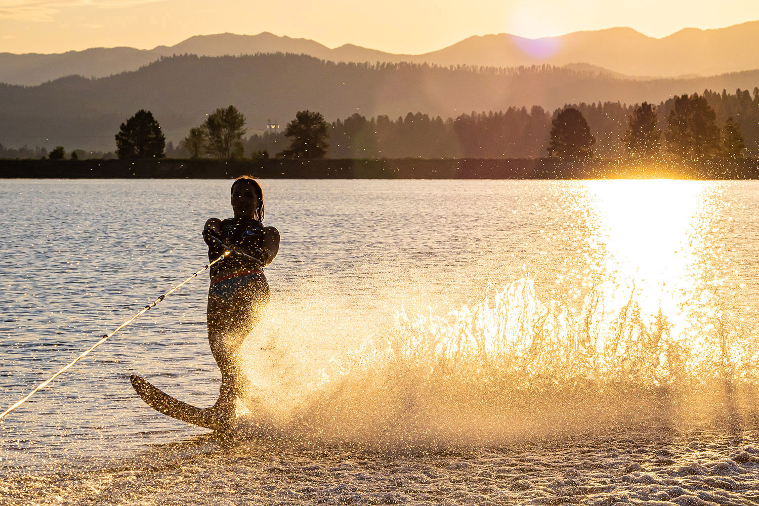 A water skier carves across a lake at sunset near Tamarack Resort in McCall, Idaho, sending up a golden spray against the warm evening light.